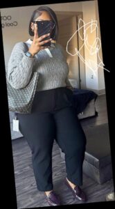 A plus size woman in

Grey cable-knit sweater with white collar detail

Black high-waisted trousers

Deep purple/burgundy patent leather loafers

Black and grey patterned tote bag

Smartwatch and black-rimmed glasses