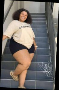 A plus size woman wearing Tan "ESSENTIALS" oversized t-shirt Black high-waist bike shorts Tan foam runners (Yeezy style) Black headband White scrunchie