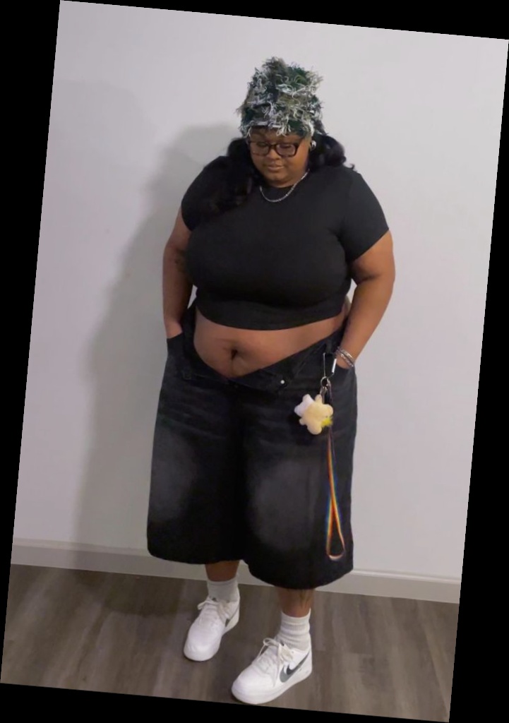 A plus size woman wearing Textured green and white fuzzy beanie (camouflage style)

Black short sleeve crewneck top

Oversized black denim bermuda shorts

White high-top sneakers (Nike Dunk style)

White socks

Chain necklace