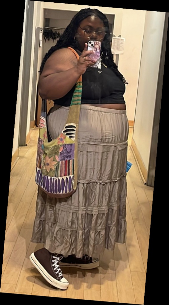 A plus size woman wearing Black short sleeve crewneck top

Brown multi-tiered maxi skirt with lace trim

Dark brown high-top sneakers (Converse style)

Oversized patchwork cross-body bag

Gold cross pendant necklace
