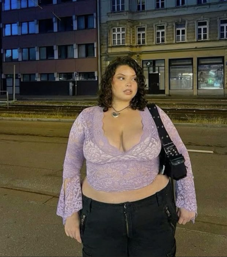 A plus size woman wearing Lavender sheer floral lace long sleeve crop top with bell sleeves Black cargo pants Black shoulder bag with stud details Silver heart pendant necklace Chain necklace