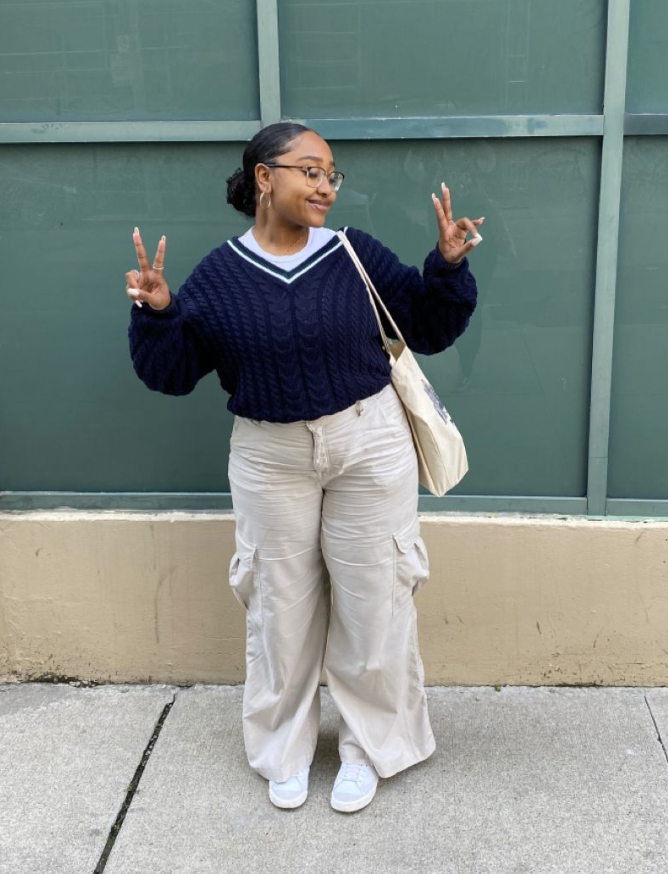 A plus size woman wearing Navy Blue Cable Knit V-Neck Sweater with white trim on the neckline White Crewneck T-shirt Beige/Light Tan Cargo Pants with large pockets and drawstring waist Light-colored (possibly white or light blue) Sneakers Small Hoop Earrings Glasses (tortoiseshell frame) Beige Tote Bag