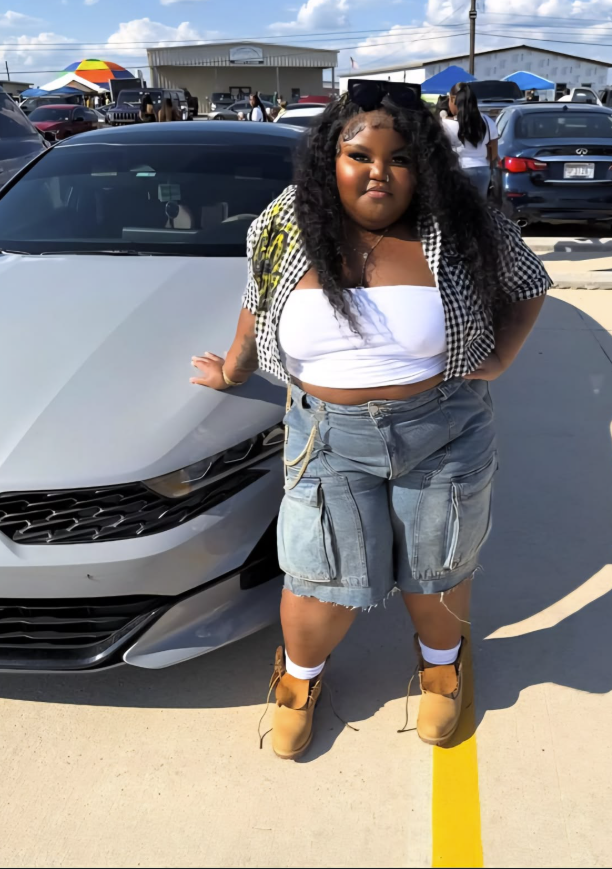 A plus size woman wearing Black and White Gingham Button-Up Shirt, short-sleeved White Tube Top/Strapless Top Long, Wide-Leg Light Wash Denim Cargo Shorts with large pockets and raw hem Beige Suede Lace-Up Boots (looks like Timberlands) White Socks Black Sunglasses (resting on head) Necklace (looks like a delicate chain with a small pendant) Bracelet (gold or silver chain) Belly Chain (gold or silver chain)