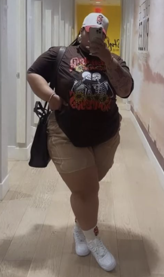 A plus size woman wearing Dark Grey/Charcoal Graphic T-Shirt, short-sleeved, featuring two skeletons holding flowers with floral accents Beige/Tan Biker Shorts White Sneakers White Socks with a small logo (looks like a red 'B' or 'R') White Baseball Cap with a red and blue logo (looks like a stylized 'B' or 'R') Black Tote Bag Bracelet (looks like a beaded stack) Ring
