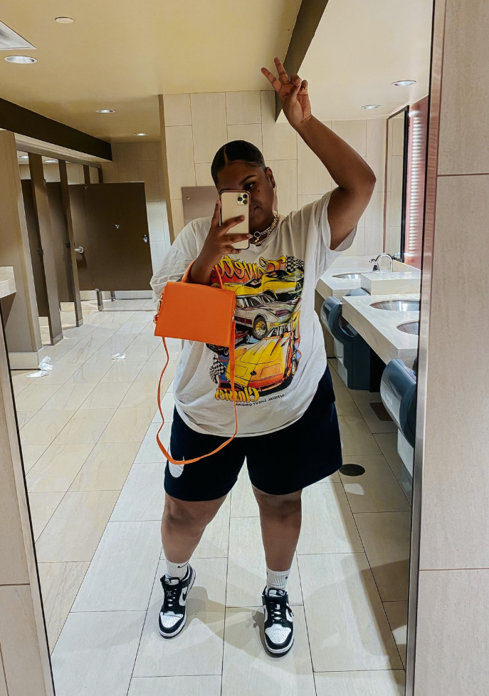 A Plus size woman wearing White Graphic T-Shirt, short-sleeved, featuring vintage cars and the word "Classics" Dark Navy/Black Athletic Shorts (sweat-short material, with a small logo) White Sneakers with black details (looks like Nike Dunks) White Socks Small Orange Square Crossbody Bag Necklace (appears to be a layered or simple chain) Gold Watch/Bracelet