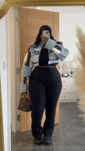 A plus size woman wearing Cropped Bomber Jacket, light blue with brown and white patches/graphics (looks like a racing-style jacket) Black Tank Top/Cami Black Flared/Wide-Leg Leggings Dark Green/Brown Suede-like Sneakers Black Baseball Cap Brown Leather Handbag with a repeating pattern (looks like a designer logo)
