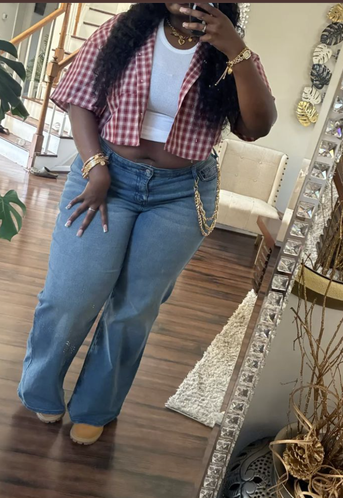 A plus size woman wearing Red and White Gingham Cropped Short-Sleeved Button-Up Shirt White Tank Top/Cami (cropped, peeking out) Light Wash Denim, Wide-Leg/Flared Jeans Tan Suede Lace-Up Boots (looks like Timberlands) Layered Gold Necklaces Stack of Bracelets (gold and possibly pearl or stone) Gold Chain Belt/Belly Chain (draping over her hip)