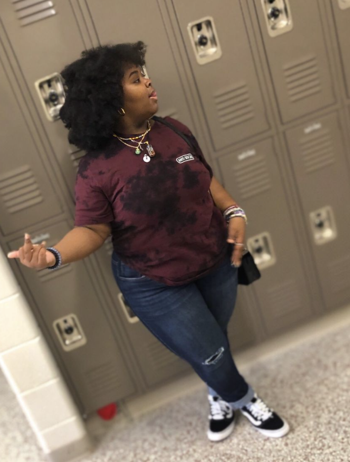 A plus size woman wearing Dark Burgundy/Maroon and Black Tie-Dye T-Shirt, short-sleeved (with a small logo on the chest) Medium Wash Distressed Skinny/Straight Leg Jeans with rips at the knees Black and White Sneakers with a star detail (looks like Golden Goose) Layered Necklaces (appears to be a mix of chain and possible natural stones) Stack of Bracelets (on both wrists) Hoops Earrings Black Handbag (small, crossbody)