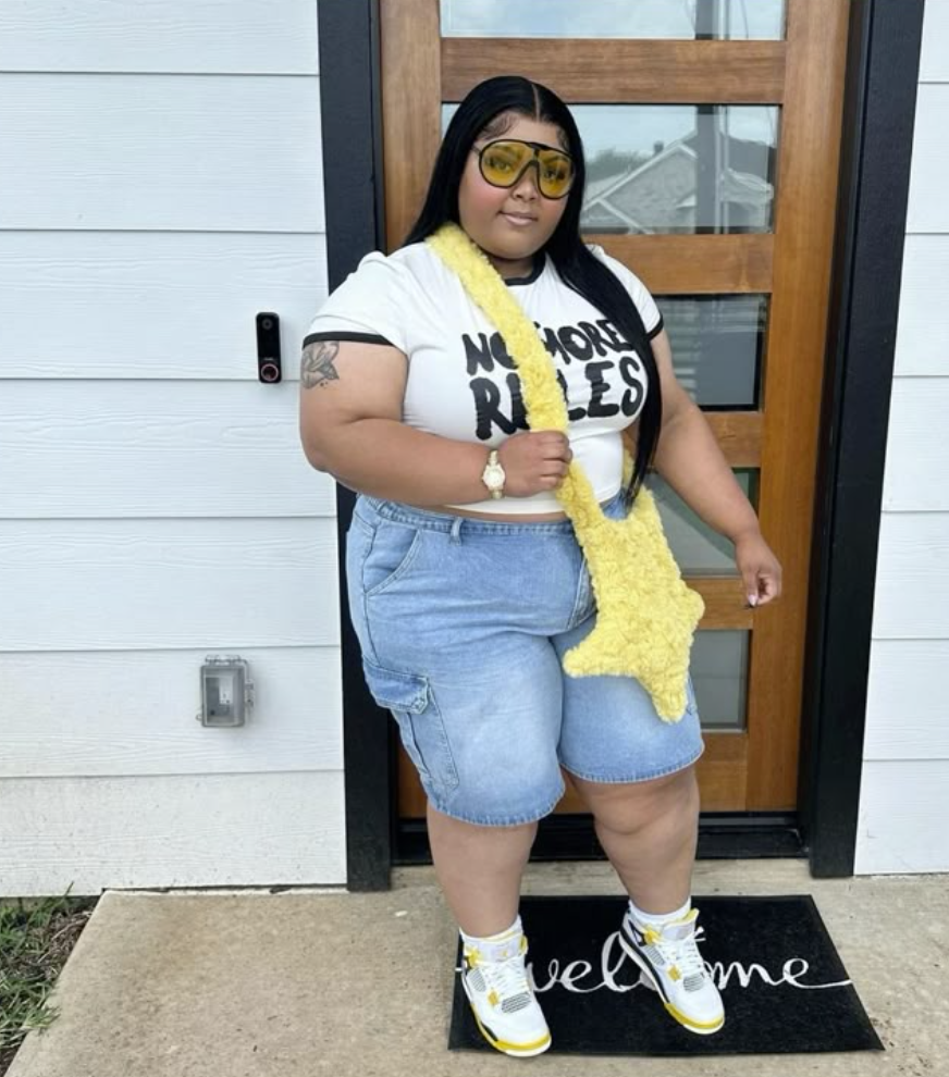 A plus size woman wearing White Cropped Ringer T-Shirt with black trim and a black and yellow graphic text Light Wash Denim Cargo Shorts with large pockets and raw hem White and Yellow High-Top Sneakers (looks like Nike Dunks or Jordans) White Socks Large Yellow Plush Star-Shaped Bag Yellow-Tinted Aviator-Style Sunglasses Layered Gold Necklaces Gold Watch