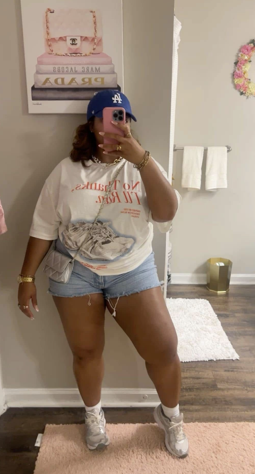 A plus size woman wearing White Graphic T-Shirt, short-sleeved, with red graphic text and a sneaker design Light Wash Denim Shorts with raw hem White Sneakers White Socks Blue Baseball Cap with white logo (looks like a sports team, possibly LA Dodgers) Small Metallic Silver Quilted Crossbody Bag Gold Chain Necklace Gold Watch Gold and Gemstone Ring