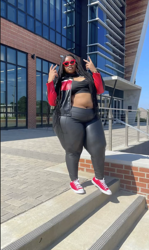 A plus size woman wearing Black scoop-neck sports bra crop top
Black coated faux-leather high-waisted leggings
Black faux-leather racer jacket with red and white color blocking
Red low-top canvas sneakers with white laces
Red thick-frame cat-eye sunglasses