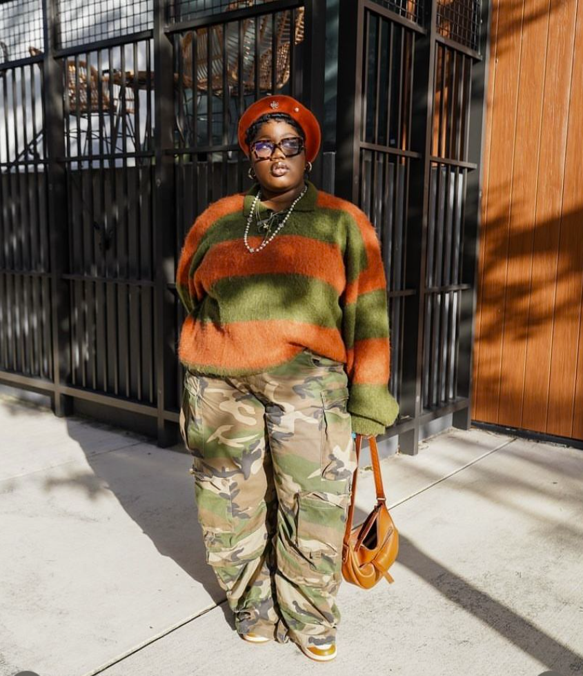 A plus size woman wearing Green and orange wide-striped fuzzy polo sweater
Camouflage wide-leg cargo pants
Burnt orange wool beret hat
Brown leather crescent shoulder bag
White and yellow chunky sneakers
Silver pearl chain necklace
