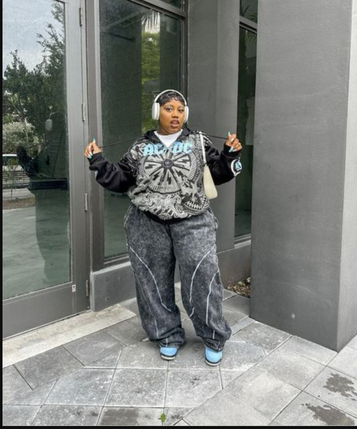 A plus size woman wearing Black and grey graphic print zip-up hoodie
Acid wash dark grey wide-leg joggers with white piping
Light blue skate sneakers
White over-ear wireless headphones
Cream leather shoulder bag
