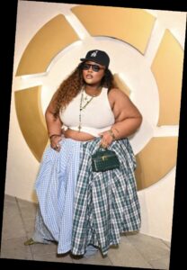A plus size woman wearing White sleeveless ribbed tank top, knotted at the waist Oversized mixed-print plaid skirt (blue gingham and green tartan) Loose fit blue jeans underneath the skirt Dark blue baseball cap (LA logo) Small square dark green quilted handbag Black sunglasses Layered gold chain necklace