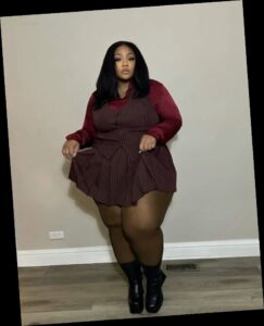 A plus size woman wearing Burgundy satin long sleeve button-down shirt Burgundy pinstripe vest and pleated mini skirt set Sheer black tights Black platform ankle boots Gold pendant necklace