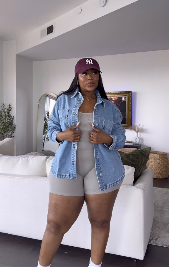 Sun-Ready Summer Vacation Outfits for Plus Size Women 5 A plus size woman wearing Light wash oversized denim button-up shirt Taupe grey ribbed knit sleeveless romper / biker short bodysuit Burgundy New York Yankees baseball cap Gold delicate pendant necklaces White crew socks