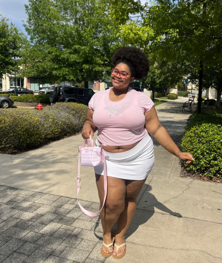 Sun-Ready Summer Vacation Outfits for Plus Size Women 6 A plus size wearing Light pink fitted short-sleeve t-shirt with a subtle graphic White low-rise mini skort / athletic skirt Pink and white gingham mini crossbody tote bag Beige thong sandals / flip flops Red-framed retro eyeglasses Silver chain bracelet