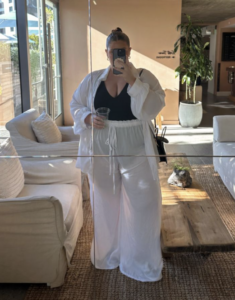 A plus size woman wearing White oversized linen button-up shirt White sheer wide-leg palazzo pants with a drawstring waist Black low-cut tank top / one-piece swimsuit Black leather shoulder bag Gold hoop earrings