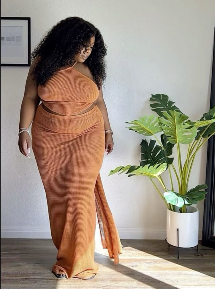 A plus size woman wearing Burnt orange waffle-knit halter neck crop top Burnt orange matching high-waisted textured maxi skirt with a side-drape train Silver chain link bracelet Gold watch with a metallic link band Black minimal slide sandals