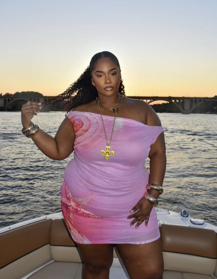 A plus size woman wearing Pink and orange starfish print mesh off-the-shoulder mini dress Chunky gold chain-link choker necklace Long gold chain necklace with a large yellow gemstone cross pendant Thick stacked gold bangle bracelets Gold statement cocktail rings