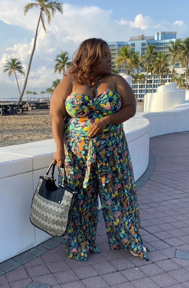 A plus size woman wearing Dark teal tropical botanical print sweetheart neckline strapless bodice with halter string Matching printed wide-leg palazzo pants with a front tie-waist Black and white geometric woven patterned tote bag with black leather top handles White minimal low-heel sandals