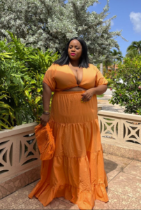A plus size woman wearing Bright orange short-sleeve crop top with a twist-front plunge neckline Bright orange matching tiered high-waisted maxi skirt with a ruffled hem Orange textured woven knit clutch bag Stacked metallic bangles / bracelets