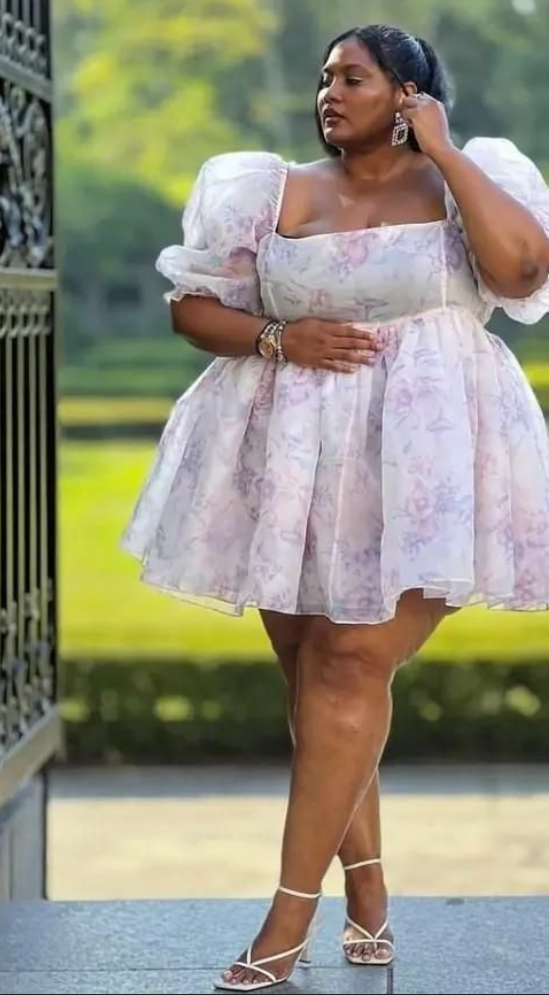 A plus size woman wearing Lavender and pastel pink floral print organza mini babydoll dress
Square neckline with sheer short puff sleeves
White strappy lace-up mid-heel sandals
Oversized square crystal statement drop earrings
Gold luxury watch with a metallic link band
Stacked diamond rhinestone chain bracelets