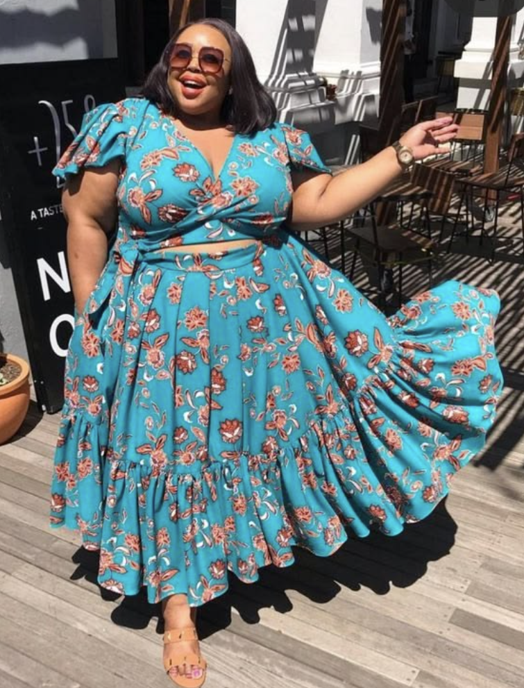 A plus size woman wearing Teal blue floral print short-sleeve wrap crop top
Teal blue matching high-waisted tiered maxi skirt with a ruffled hem
Tan leather slide sandals with a thick strap
Oversized tortoiseshell sunglasses
Gold luxury watch with a link band