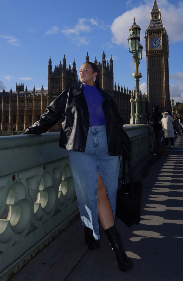A plus size woman wearing Blue mock-neck lace mesh top Black faux leather moto jacket High-waisted medium wash denim maxi skirt with a front slit Black platform leather ankle boots Silver statement floral-shaped earrings Large black leather tote bag 