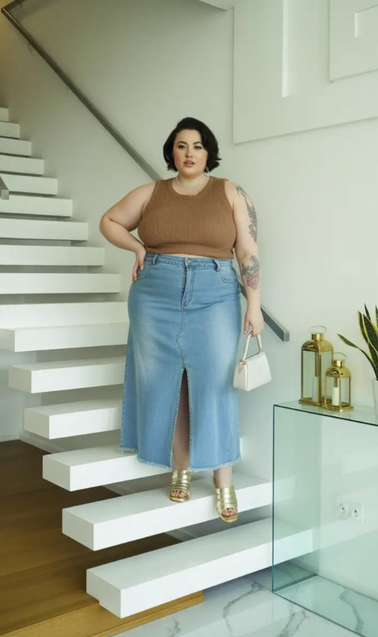 A plus size woman wearing Brown ribbed knit cropped tank top Light wash denim maxi skirt with a frayed hem and front slit Gold metallic multi-strap slide sandals Small white leather top-handle handbag Classic white pearl strand necklace