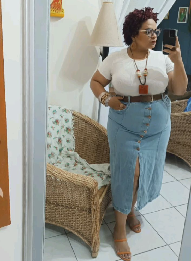 A plus size woman wearing White ribbed short-sleeve t-shirt Light wash denim midi skirt with front button closures and a slit Thick brown leather waist belt with a silver buckle Orange faux leather slide sandals Chunky statement necklace with geometric resin beads Clear-framed square eyeglasses Stacked neutral beaded bracelets