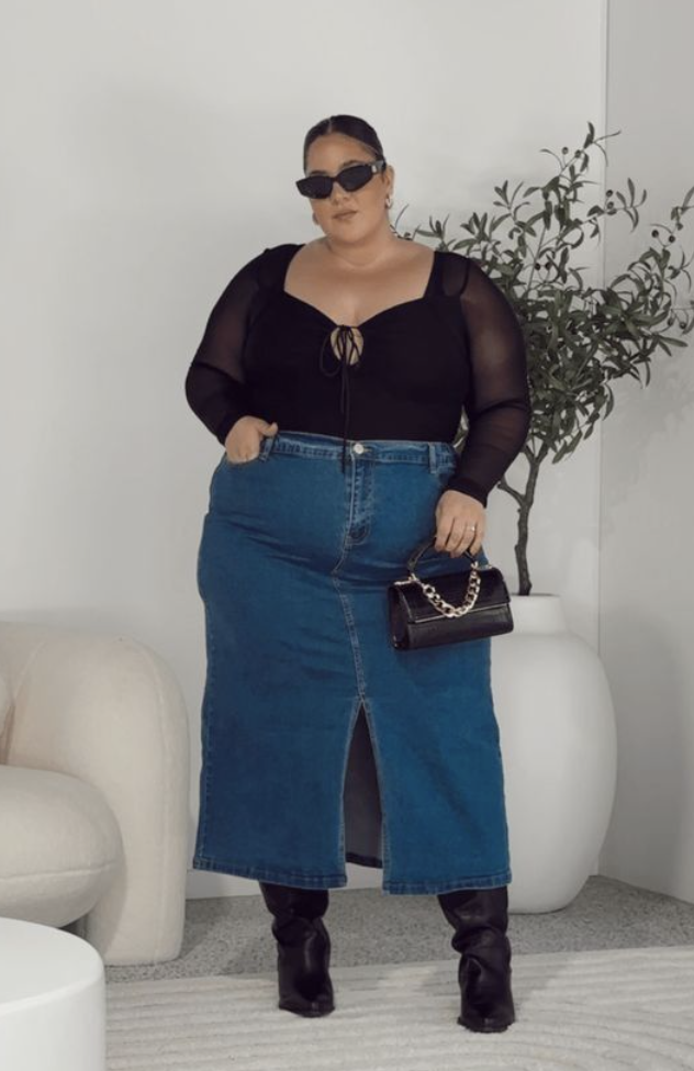 A plus size woman wearing Black sweetheart neckline long-sleeve mesh panel top with front tie detail Medium wash denim maxi skirt with a front slit Black leather knee-high boots Small black alligator texture handbag with a chunky silver chain strap Black rectangular sunglasses