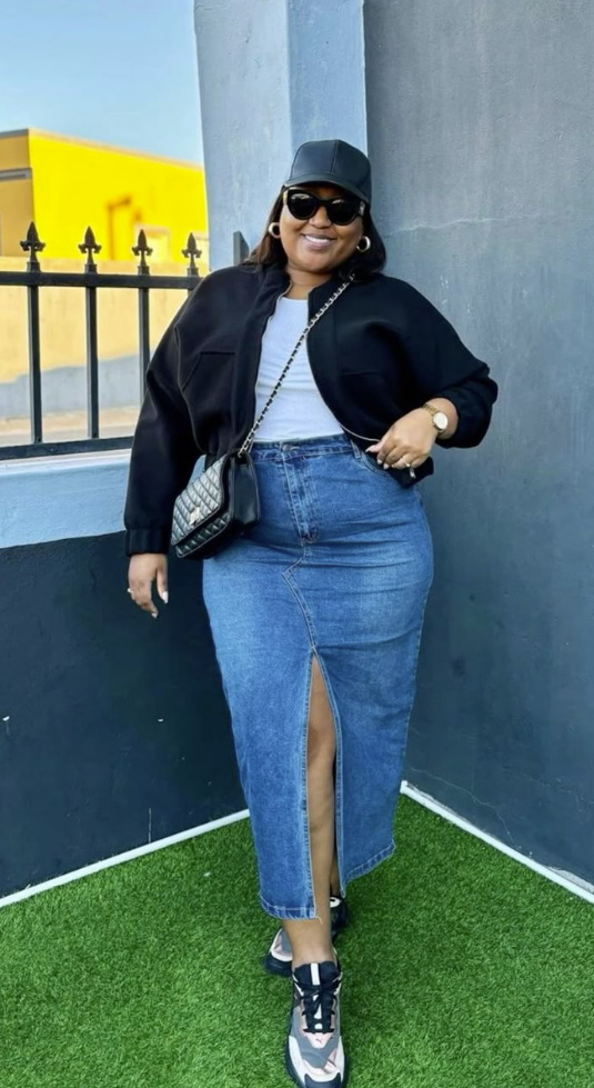 A plus size woman wearing Black oversized cropped bomber jacket White classic crewneck t-shirt Medium wash high-waisted denim maxi skirt with a front slit Black and white athletic panel sneakers Black quilted leather crossbody bag with a gold chain strap Black leather baseball cap Black round sunglasses