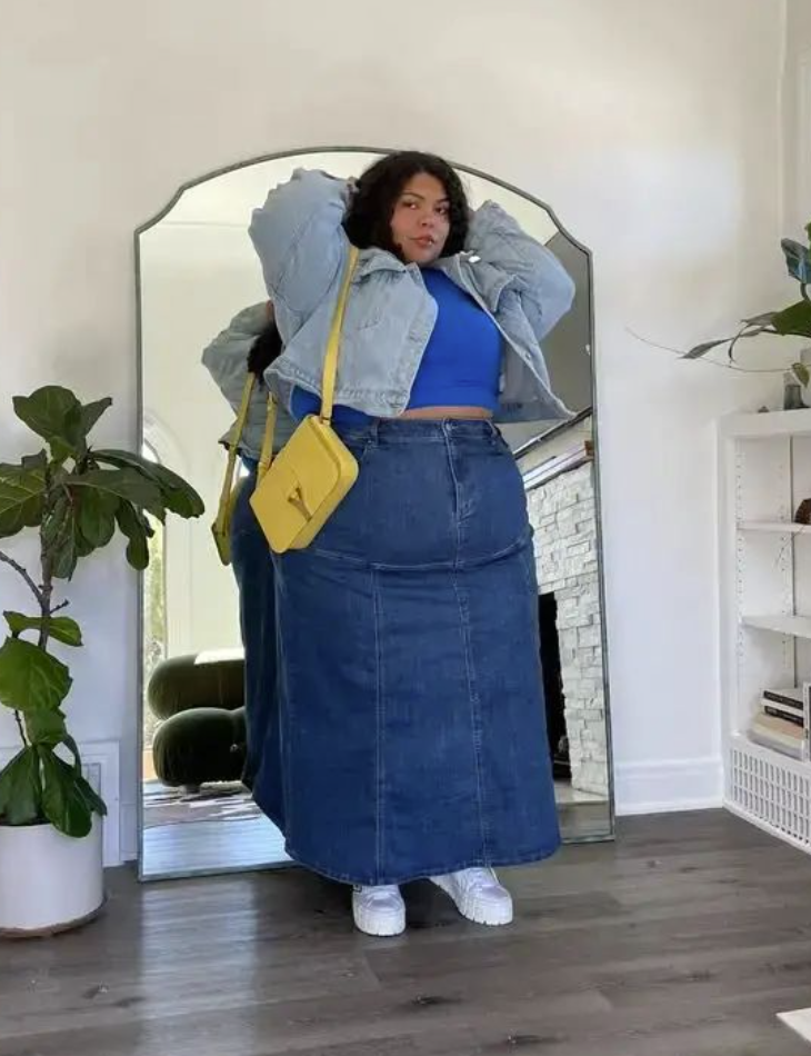 A plus size woman wearing Light wash oversized denim jacket Royal blue cropped tank top Dark wash denim maxi skirt with panel stitching White platform leather sneakers Bright yellow leather shoulder bag with a top flap