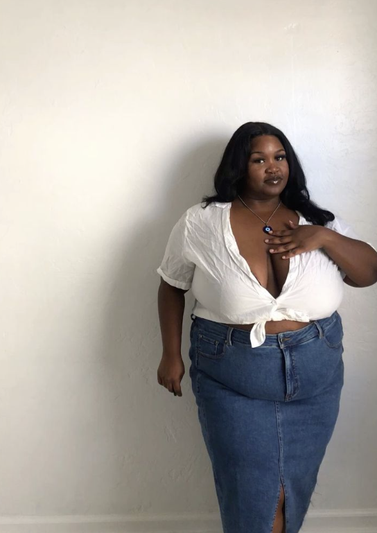 A plus size woman who is wearing White short-sleeve button-down shirt tied at the waist Medium wash denim maxi skirt with a front slit Silver chain necklace with a blue evil eye pendant