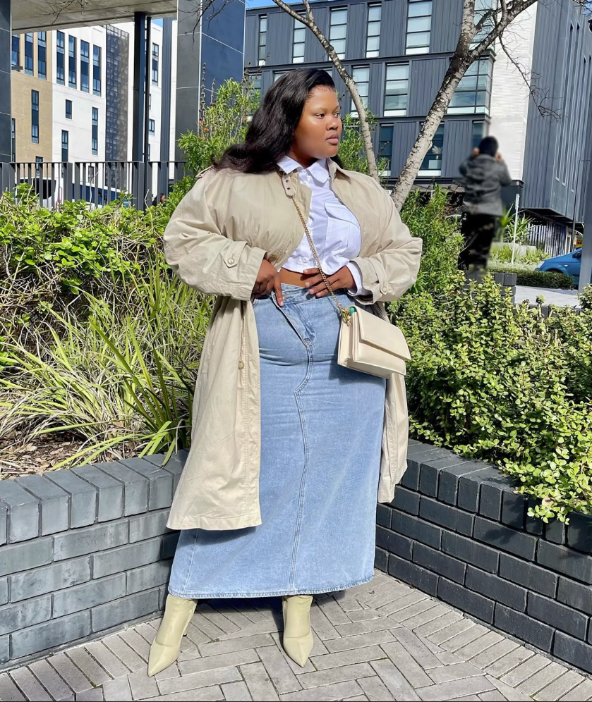 A plus size woman wearing Lightweight beige duster trench coat White collared button-up crop shirt Light wash high-waisted denim maxi skirt Cream pointed-toe heeled ankle boots Cream leather crossbody bag with a gold chain strap
