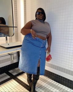 A plus size woman wearing Taupe sleeveless cable-knit turtleneck crop sweater Medium wash denim maxi skirt with a front slit Black leather over-the-knee boots Small red leather structured bag with gold hardware Black thick-frame rectangular sunglasses Silver hoop earrings