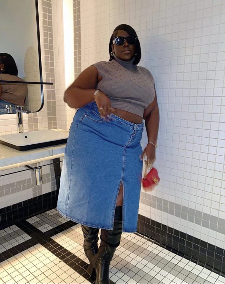 A plus size woman wearing Taupe sleeveless cable-knit turtleneck crop sweater Medium wash denim maxi skirt with a front slit Black leather over-the-knee boots Small red leather structured bag with gold hardware Black thick-frame rectangular sunglasses Silver hoop earrings