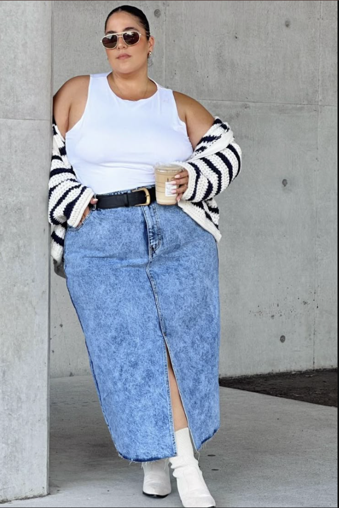 A plus size woman wearing Black and white striped chunky knit open-front cardigan White scoop-neck tank top Black leather belt with a square gold buckle Acid wash marbled denim maxi skirt with a frayed hem and front slit White leather pointed-toe ankle boots Gold wire aviator sunglasses
