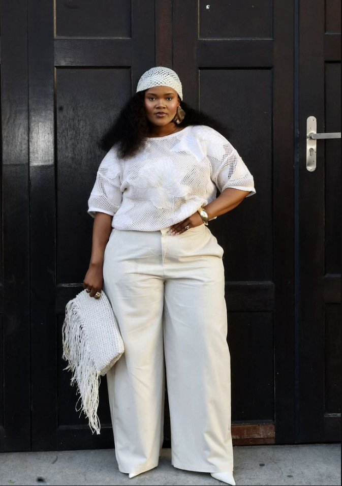 A plus size woman wearing White short-sleeve open-knit crochet sweater with floral patterns Off-white wide-leg tailored trousers White leather pointed-toe heeled boots Cream crochet knit beanie hat White woven leather fringe clutch handbag Gold statement teardrop earrings Gold chunky bangle bracelet