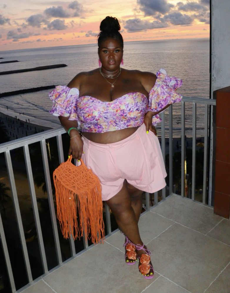 Beach Queen Energy: Stylish Outfits for Plus Size Women by the Sea 9 A plus size woman wearing Pink and purple floral print off-the-shoulder crop top with tiered ruffle sleeves
Light pink high-waisted wide-leg shorts
Bright orange woven macrame tote bag with dramatic long fringe tassels
Pink square-toe strappy sandals adorned with large orange fabric rosettes
Hot pink statement drop earrings
Layered gold chain necklaces
Green resin bangle bracelet