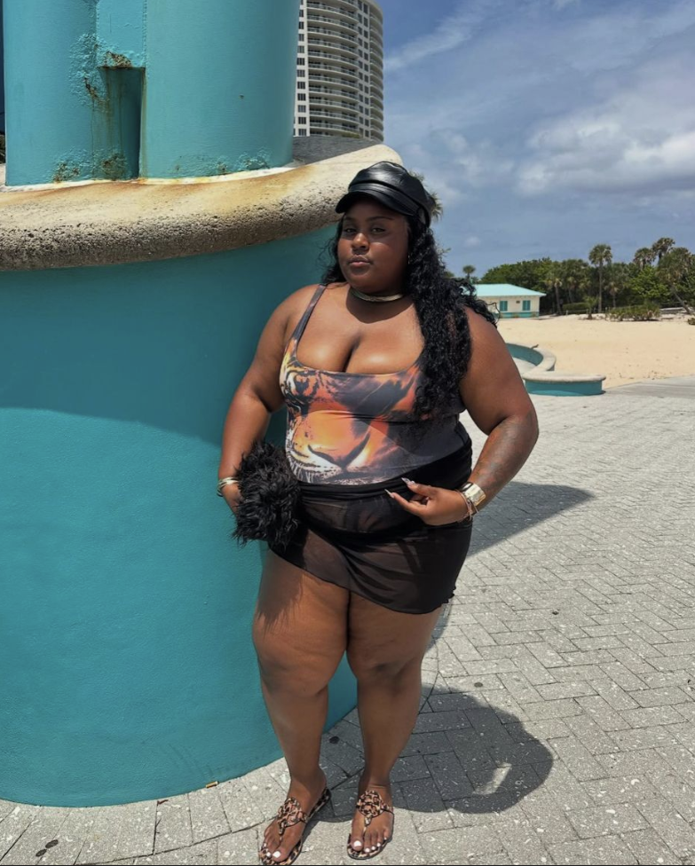 Beach Queen Energy: Stylish Outfits for Plus Size Women by the Sea 10 A plus size woman wearing Orange, black, and white tiger face graphic print one-piece swimsuit
Black sheer mesh mini skirt cover-up
Black faux-leather captain baker boy hat
Leopard print thong sandals
Black furry textured clutch bag
Chunky silver collar necklace
Wide silver cuff bracelets