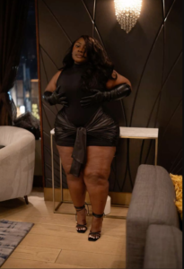 A plus size woman wearing Black sleeveless mock-neck bodycon mini dress Black satin wrap-around waist tie sash Black leather long opera gloves Black strappy square-toe stiletto sandals Crystal drop earrings