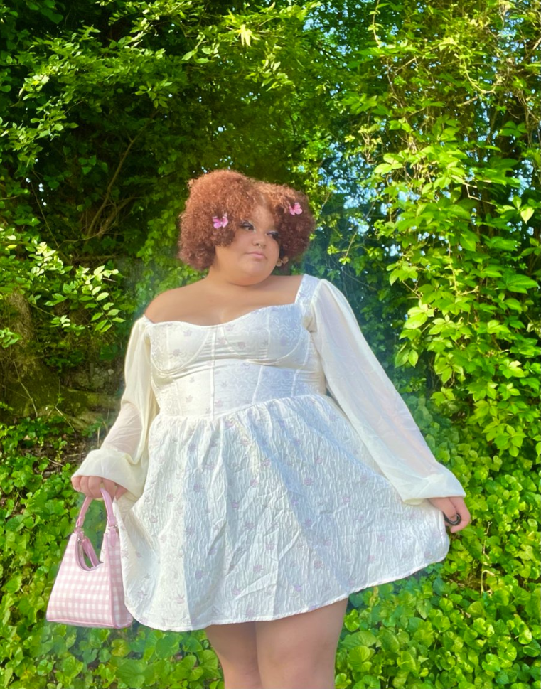A plus size woman wearing Cream jacquard floral mini dress with corset bodice
Sheer cream long bell sleeves
Pink butterfly hair clips
Pink gingham shoulder bag