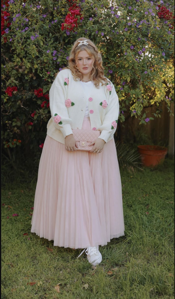 A plus size woman wearing Cream knit cardigan with 3D pink rose appliqués
White crew neck t-shirt
Blush pink pleated tulle maxi skirt
Pink quilted crossbody bag with gold chain strap
White satin hair bow clip
White lace-up sneakers