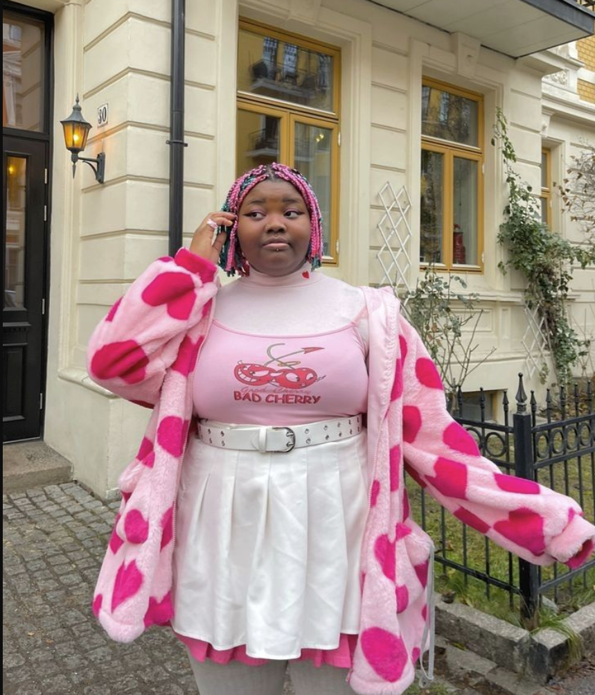 A plus size woman wearing Pink and red heart print faux fur coat
Pink cherry graphic tank top
Light pink turtleneck undershirt
White pleated mini skirt
White double-grommet faux leather belt
Pink heart statement ring