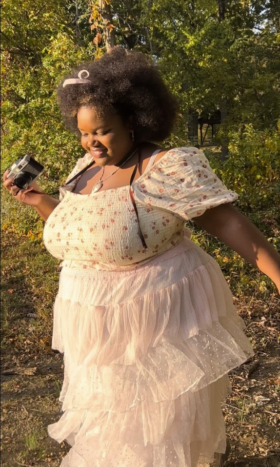 A plus size woman wearing Cream and pink floral print smocked short sleeve top
Cream tiered tulle ruffle maxi skirt
Pink ribbon hair bow
Silver heart pendant necklace