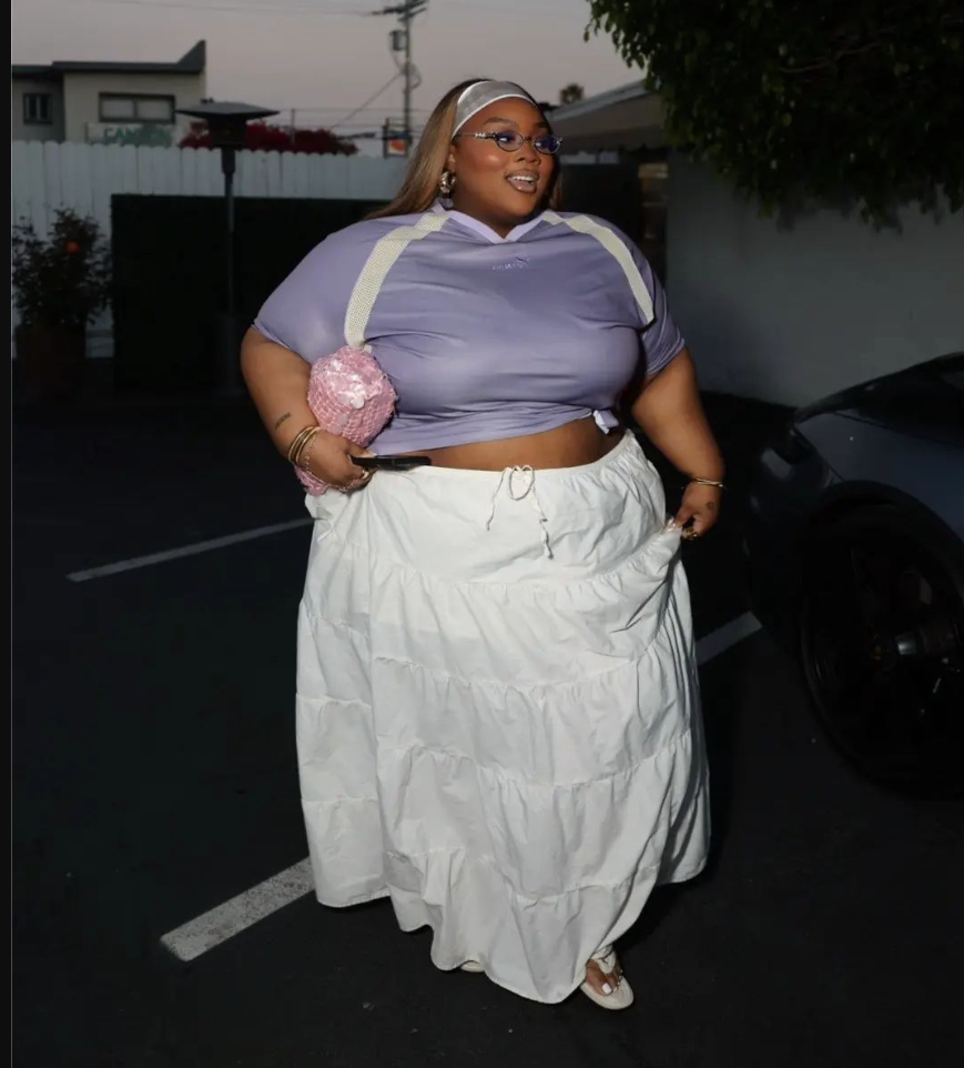 Skirt Season Forever: Flattering Skirt Outfits for Plus Size Women 2 A plus size woman wearing Lavender cropped hoodie-style top
White suspender straps
White tiered ruffled maxi skirt
Pink sequined handbag
Silver headband
Blue-tinted sunglasses
Gold hoop earrings
Gold bangles
White slide sandals