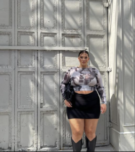 A plus size woman wearing Black and white patterned mesh long-sleeve crop top Black leather-look mini skirt Black knee-high cowboy boots Silver cross necklace Black shoulder bag