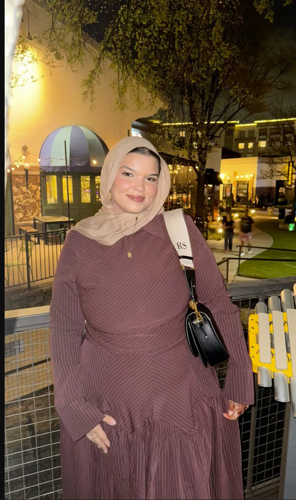 A plus size woman wearing Beige hijab Mauve pinstriped long-sleeve tiered dress Black crossbody handbag with white strap Gold pendant necklace Gold earrings